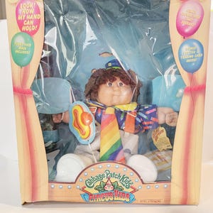 May include: A Cabbage Patch Kids Circus King doll in a blue and white clown costume with a rainbow tie and a hot dog balloon. The doll is in a cardboard box with the words "Cabbage Patch Kids Circus King" on the front.