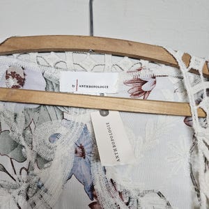 Op de afbeelding: Een wit kledingstuk met bloemenpatroon en kanten rand, hangend aan een houten hanger. Het kledingstuk heeft een label met de tekst "by ANTHROPOLOGIE". Het bloemenpatroon bevat blauwe en bruine bloemen.