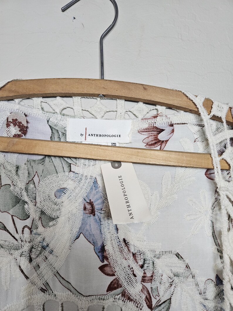 Op de afbeelding: Een wit kledingstuk met bloemenpatroon en kanten rand, hangend aan een houten hanger. Het kledingstuk heeft een label met de tekst "by ANTHROPOLOGIE". Het bloemenpatroon bevat blauwe en bruine bloemen.