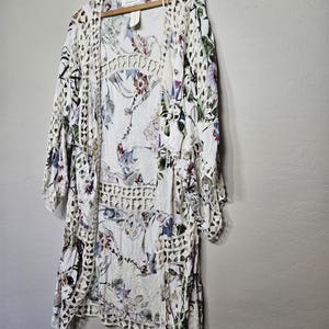 Op de afbeelding: Een witte jurk met bloemenprint en een open geweven gehaakt vest hangen aan een houten hanger. De jurk heeft een delicaat bloemenpatroon met paarse, blauwe en rode accenten. Het vest heeft een geometrisch open geweven ontwerp.