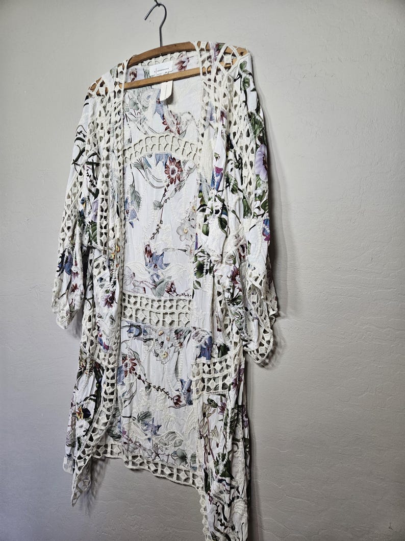 Op de afbeelding: Een witte jurk met bloemenprint en een open geweven gehaakt vest hangen aan een houten hanger. De jurk heeft een delicaat bloemenpatroon met paarse, blauwe en rode accenten. Het vest heeft een geometrisch open geweven ontwerp.
