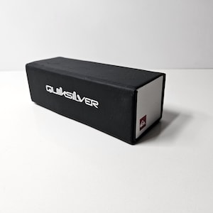 Może przedstawiać: Prostokątne czarne pudełko do przechowywania marki Quiksilver o matowym wykończeniu. Nazwa marki Quiksilver jest nadrukowana na biało na górnej powierzchni. Jeden koniec pudełka posiada biały panel z małym czerwonym kwadratowym logo z ikoną.