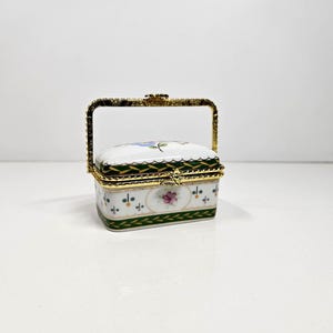 May include: A rectangular porcelain trinket box with a hinged lid and a gold-tone metal handle. The white surface features a green and gold decorative border, small floral motifs, and a central pink rose design with gold metal trim edges.