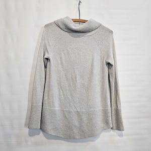 K&ouml;nnte beinhalten: Ein hellgrauer Pullover mit einem Rollkragen und langen &Auml;rmeln. Der Pullover h&auml;ngt an einem Holzb&uuml;gel vor einem wei&szlig;en Hintergrund. Der Saum und die Manschetten haben eine gerippte Textur.