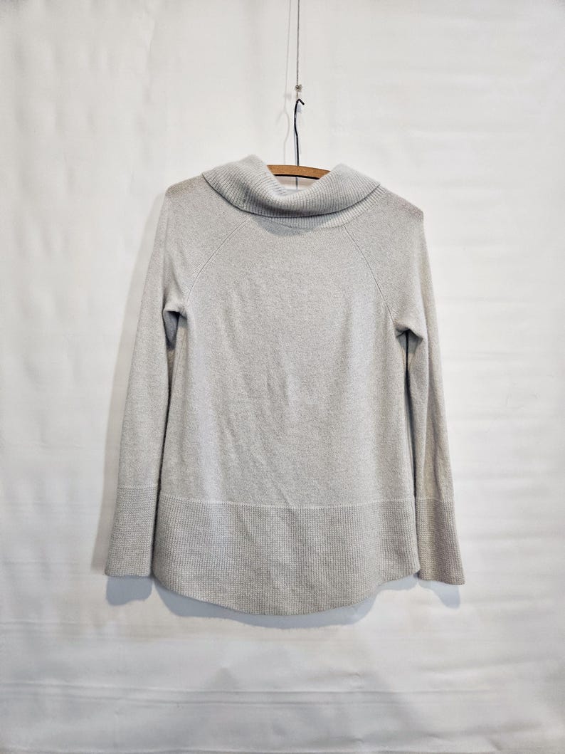 K&ouml;nnte beinhalten: Ein hellgrauer Pullover mit einem Rollkragen und langen &Auml;rmeln. Der Pullover h&auml;ngt an einem Holzb&uuml;gel vor einem wei&szlig;en Hintergrund. Der Saum und die Manschetten haben eine gerippte Textur.