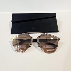 Op de afbeelding: Gouden aviator zonnebril met bruine verlopende glazen. De zonnebril zit in een zwarte etui met het woord "Dior" erop gedrukt.