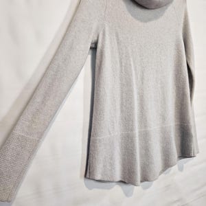 K&ouml;nnte beinhalten: Ein hellgrauer Pullover mit einem Rollkragen und langen &Auml;rmeln. Der Pullover hat einen gerippten Saum und B&uuml;ndchen. Das Kleidungsst&uuml;ck h&auml;ngt an einem Holzb&uuml;gel vor einem wei&szlig;en Hintergrund.