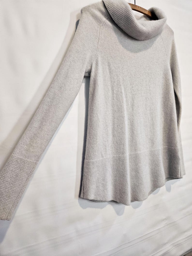 K&ouml;nnte beinhalten: Ein hellgrauer Pullover mit einem Rollkragen und langen &Auml;rmeln. Der Pullover hat einen gerippten Saum und B&uuml;ndchen. Das Kleidungsst&uuml;ck h&auml;ngt an einem Holzb&uuml;gel vor einem wei&szlig;en Hintergrund.