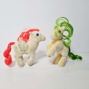 May include: Two vintage toy ponies. One is off-white with red hair and tail, and a wing design. The other is yellow with green hair. Both have a soft, plush texture and are standing on a white surface.
