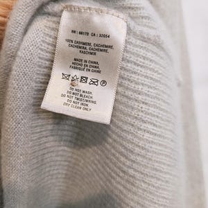 K&ouml;nnte beinhalten: Nahaufnahme eines hellblauen Kaschmirpullovers mit Etikett. Das Etikett lautet "100% CASHMERE" und "MADE IN CHINA". Pflegehinweise umfassen "DRY CLEAN ONLY". Der Pullover besteht aus einem weichen, strukturierten Material.