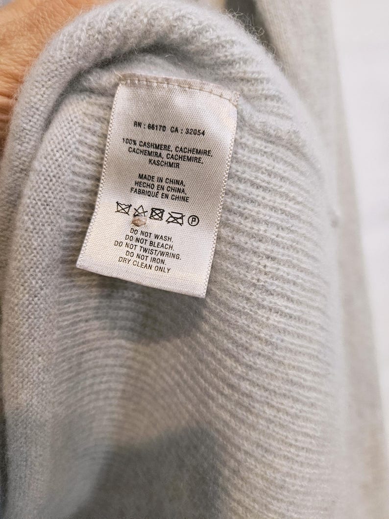 K&ouml;nnte beinhalten: Nahaufnahme eines hellblauen Kaschmirpullovers mit Etikett. Das Etikett lautet "100% CASHMERE" und "MADE IN CHINA". Pflegehinweise umfassen "DRY CLEAN ONLY". Der Pullover besteht aus einem weichen, strukturierten Material.