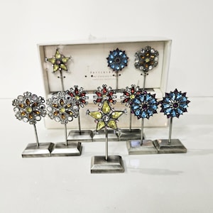Könnte beinhalten: Eine Sammlung dekorativer Ornamente in Blumen- und Sternformen mit farbigen Edelsteinakzenten. Die Ornamente sind auf silbernen Stielen und quadratischen Sockeln angebracht. Die Farbpalette umfasst Gelb, Rot, Blau und Braun. Einige sind in einer weißen Box ausgestellt.