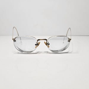 Könnte beinhalten: Eine Brille mit dünnen, goldfarbenen Metallrahmen in einem Halbrand-Design. Die Gläser sind klar und oval geformt, gestützt durch verstellbare Nasenpads und schlanke, geschwungene Bügel, die von den Rändern des Rahmens nach außen verlaufen.
