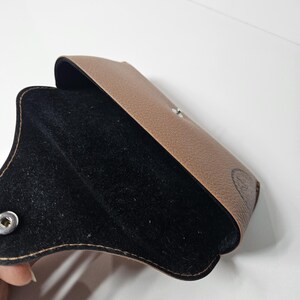 May include: A tan textured glasses case with a black interior lining. The case features a silver-colored metal snap button closure on the front flap. A small black logo is visible on the lower right side of the exterior surface of the case.
