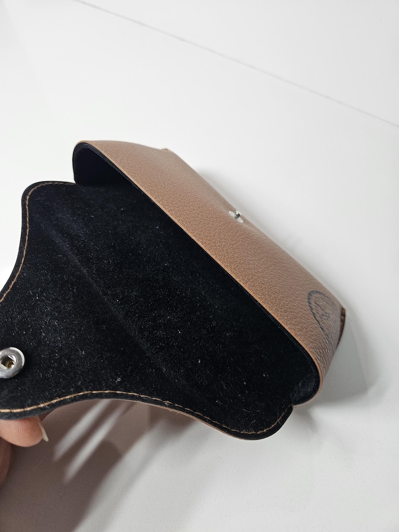 May include: A tan textured glasses case with a black interior lining. The case features a silver-colored metal snap button closure on the front flap. A small black logo is visible on the lower right side of the exterior surface of the case.