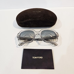 Może przedstawiać: Okulary przeciwsłoneczne z przezroczystą oprawką i szarymi szkłami, z logo Tom Ford na etui. Okulary przeciwsłoneczne leżą na czarnej powierzchni.