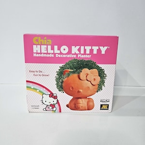 Puede incluir: Una caja rosa con las palabras "Chia HELLO KITTY Handmade Decorative Planter". La maceta es una figura de Hello Kitty de color terracota con una parte superior verde y frondosa. La caja también presenta un arcoíris y el texto "Easy to Do... Fun to Grow!"