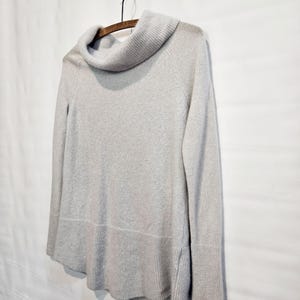 K&ouml;nnte beinhalten: Ein hellgrauer Pullover mit einem Rollkragen, langen &Auml;rmeln und gerippten B&uuml;ndchen und Saum. Der Pullover h&auml;ngt an einem Holzkleiderb&uuml;gel vor einem wei&szlig;en Hintergrund. Das Design des Pullovers ist schlicht und klassisch.