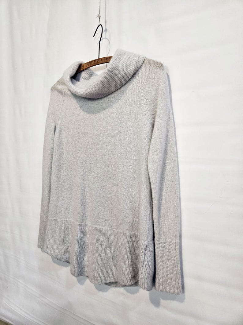 K&ouml;nnte beinhalten: Ein hellgrauer Pullover mit einem Rollkragen, langen &Auml;rmeln und gerippten B&uuml;ndchen und Saum. Der Pullover h&auml;ngt an einem Holzkleiderb&uuml;gel vor einem wei&szlig;en Hintergrund. Das Design des Pullovers ist schlicht und klassisch.