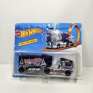 May include: A Hot Wheels toy semi-truck in its original blister packaging. The truck features a dark purple cab and trailer with light blue flame graphics and the Hot Wheels logo. The packaging includes the text Hot Wheels and Great for Track.