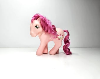 Op de afbeelding: Een lichtroze plastic pony speelgoedfiguur met fel magenta manen en een bijpassende krullende staart. De pony heeft een uitdrukking met groene ogen en een kleine afbeelding op de flank van een trosje rode aardbeien met groene blaadjes.