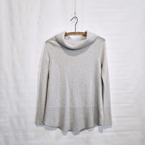 K&ouml;nnte beinhalten: Ein hellgrauer Pullover mit einem Rollkragen, der an einem Holzb&uuml;gel h&auml;ngt. Der Pullover hat lange &Auml;rmel und einen gerippten Saum. Der Hintergrund ist eine schlichte wei&szlig;e Oberfl&auml;che.