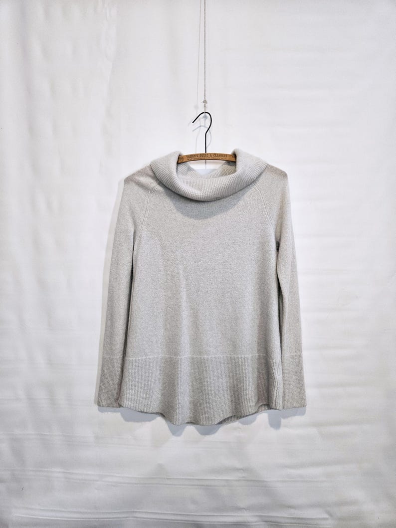 K&ouml;nnte beinhalten: Ein hellgrauer Pullover mit einem Rollkragen, der an einem Holzb&uuml;gel h&auml;ngt. Der Pullover hat lange &Auml;rmel und einen gerippten Saum. Der Hintergrund ist eine schlichte wei&szlig;e Oberfl&auml;che.