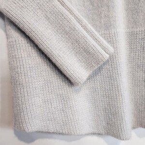 K&ouml;nnte beinhalten: Nahaufnahme eines hellgrauen Pullovers. Die Rippstrickstruktur ist an &Auml;rmel und K&ouml;rper des Kleidungsst&uuml;cks zu sehen. Der Pullover hat ein schlichtes, klassisches Design mit gerippten S&auml;umen.