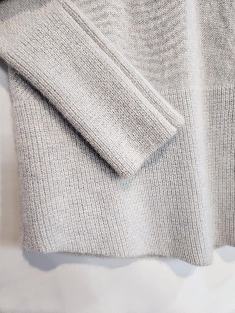 K&ouml;nnte beinhalten: Nahaufnahme eines hellgrauen Pullovers. Die Rippstrickstruktur ist an &Auml;rmel und K&ouml;rper des Kleidungsst&uuml;cks zu sehen. Der Pullover hat ein schlichtes, klassisches Design mit gerippten S&auml;umen.