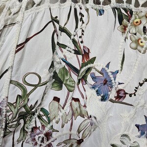 Op de afbeelding: Witte stof met een bloemenpatroon met groene bladeren en paarse, blauwe en witte bloemen. De stof heeft een decoratieve witte kanten rand en een geometrisch kanten paneel aan de bovenkant. Het algehele ontwerp is een botanisch thema.