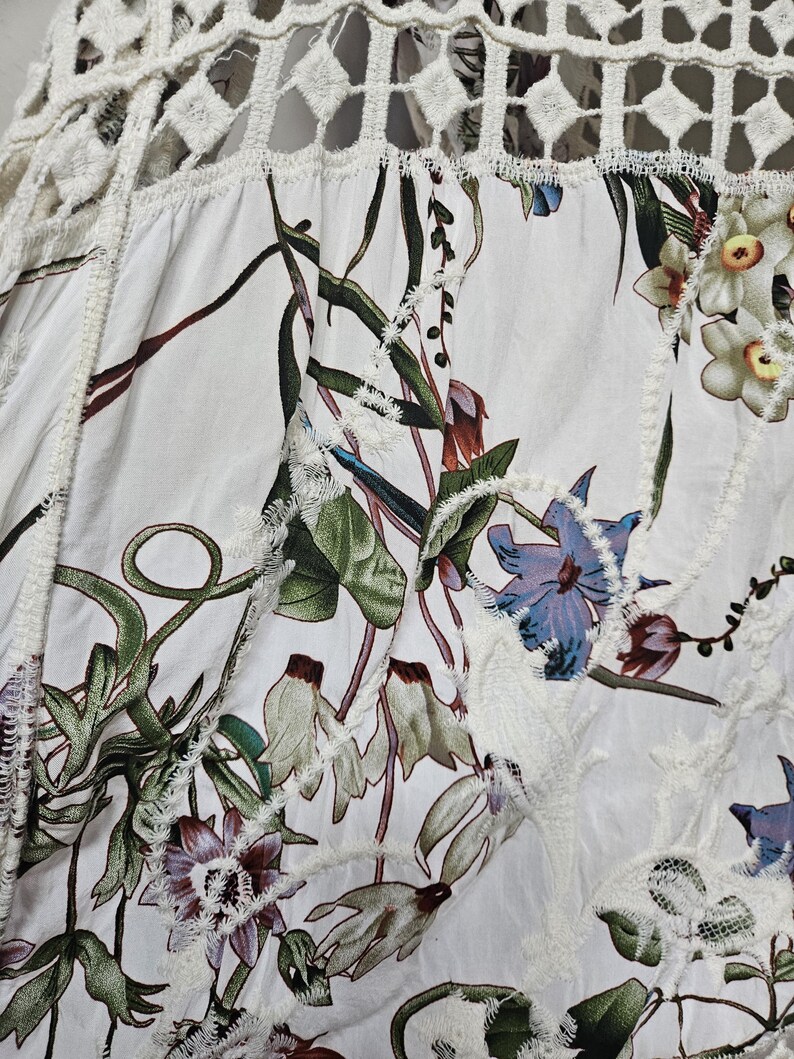 Op de afbeelding: Witte stof met een bloemenpatroon met groene bladeren en paarse, blauwe en witte bloemen. De stof heeft een decoratieve witte kanten rand en een geometrisch kanten paneel aan de bovenkant. Het algehele ontwerp is een botanisch thema.