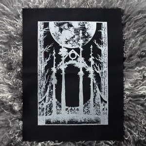 May include: A black and white art print depicting a forest scene with tall trees, a full moon, and a geometric shape. The print is on black paper with a silver design. The artwork is displayed on a grey, textured surface.