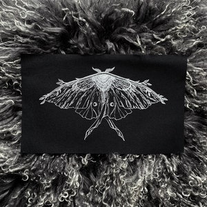 May include: A black rectangular patch featuring a detailed white illustration of a moth with outstretched wings. The moth design includes intricate wing patterns and antennae. The patch is set against a textured, gray background.