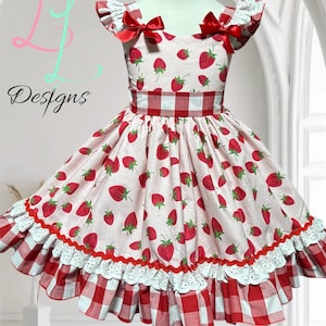 May include: A pink and white strawberry-print dress with a red gingham skirt and trim. The dress features a fitted bodice, a red gingham belt, and red bows on the shoulders. The text "L L Designs" is visible.