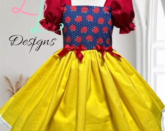 May include: A dress featuring a dark blue bodice with a white polka dot and red apple pattern, short red puff sleeves, and a solid yellow skirt. The waist is accented with two small red bows, and the hem is finished with a white lace trim.