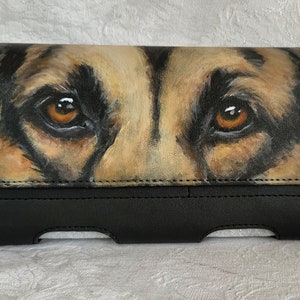 May include: A black leather phone case with a painted image of a German Shepherd's face. The dog's eyes are brown and the fur is a mix of brown and tan.