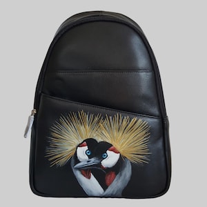 May include: Black backpack featuring a painted crowned crane design. The crane has a yellow crown, blue eyes, and red and white markings. The bag has a zippered main compartment and a front pocket.