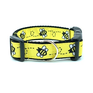 May include: A yellow dog collar with black bees and a black buckle. The collar has a silver D-ring.