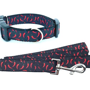 May include: A black dog collar and leash set with a red chili pepper pattern. The collar has a black plastic buckle and a silver D-ring. The leash has a silver clip.