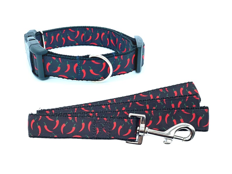 May include: A black dog collar and leash set with a red chili pepper pattern. The collar has a black plastic buckle and a silver D-ring. The leash has a silver clip.