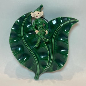 May include: A ceramic decorative piece featuring a seated elf figure atop a large, glossy green leaf. The elf is dressed in a green outfit with a matching hat. The leaf-shaped dish has detailed veining.