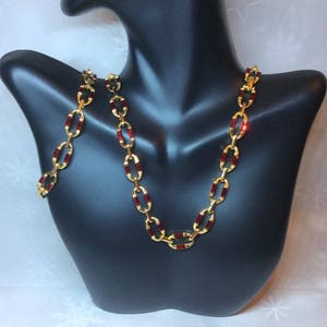 May include: A gold chain necklace and bracelet set with red rectangular stones. The necklace and bracelet are on a black mannequin.