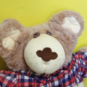 May include: A plush teddy bear with a light brown fur and a cream-colored face. The bear has brown eyes and a dark brown nose. It is wearing a red, white, and blue plaid shirt. The background is yellow.