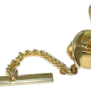 May include: A gold-colored tie tack with a chain and a bar. The tie tack has a round, flat face with a decorative edge and a small, hinged clasp. The chain connects to a rectangular bar.