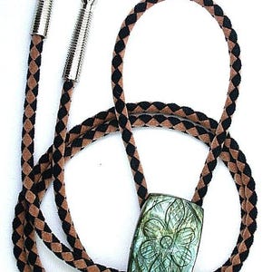 May include: A bolo tie with a braided black and brown cord. The tie features a rectangular pendant with a carved floral design in shades of green and blue. The cord ends are silver.
