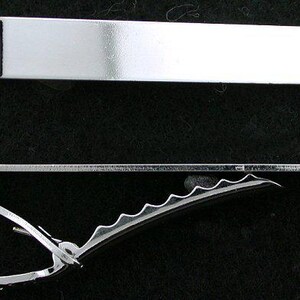 May include: A shiny silver tie bar and clip, displayed against a black background. The tie bar is rectangular, while the clip has a curved design with a serrated edge. The metal reflects light, highlighting its polished finish.