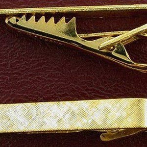 May include: Two gold-colored tie clips are displayed against a dark red background. The top clip has a serrated edge, while the bottom clip features a textured, rectangular design. These accessories are designed to secure a necktie.