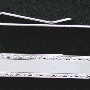 May include: Two silver-colored tie clips. One is a simple, curved bar clip. The other is a rectangular clip with a decorative border. Both clips are displayed against a dark background.