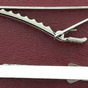 May include: Two silver-toned tie clips are displayed against a textured, burgundy background. The top clip has a serrated edge, while the bottom clip is a simple, rectangular design. These accessories are classic additions to any wardrobe.
