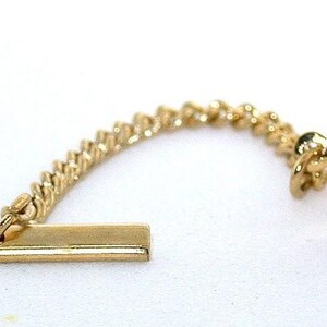 May include: A gold tie tack featuring a round setting with a faceted, vibrant pink gemstone at the center. The circular head is connected by a short, delicate gold chain to a horizontal gold bar, which serves as the fastening mechanism for the piece.